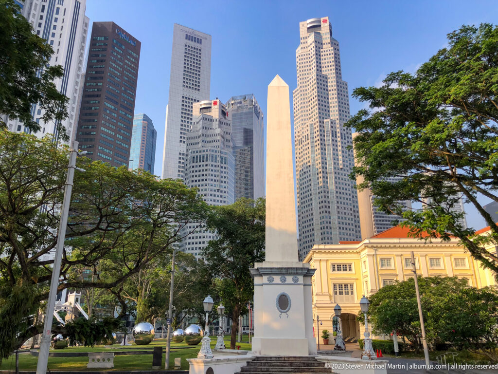 Slender pale stone obelisk in Empress Place with skyscrapers behind.