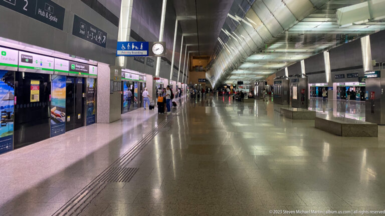 Wide interior of a Singapore MRT station with platform doors, signs, and waiting passengers.