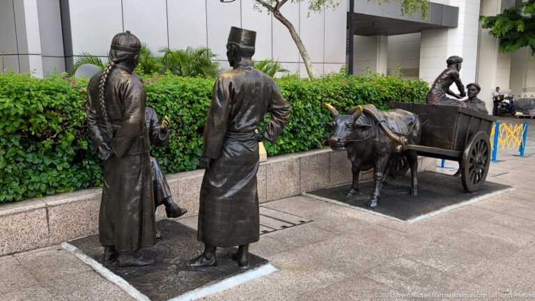 Bronze figures standing beside a buffalo cart on a landscaped riverside walkway.