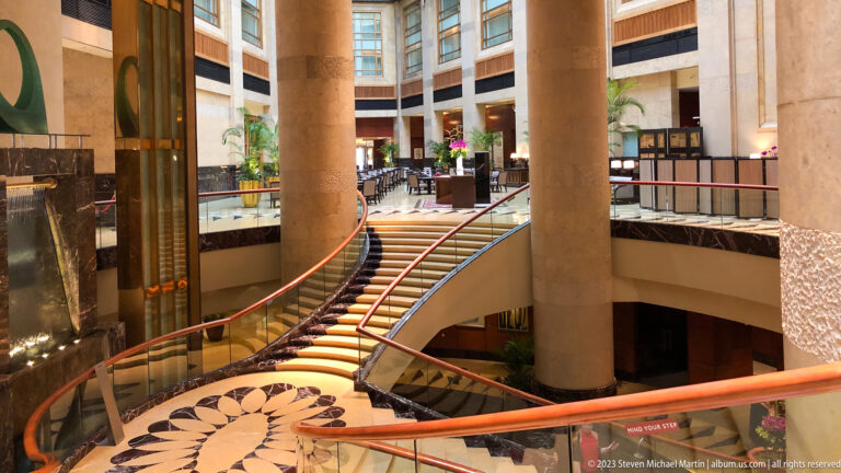 Curved carpeted staircase with red handrail in a marble hotel atrium.