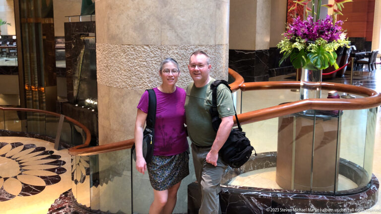 Two adult travelers standing together beside a curved railing in a hotel atrium.
