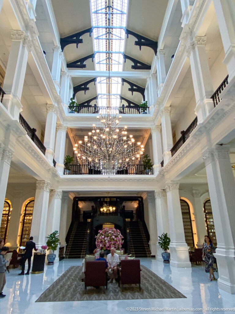 Wide view of Raffles Hotel lobby with white galleries, chandelier, seating, and dark stairs.