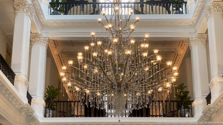 Large branching chandelier with crystal drops between white columns and an upper balcony.