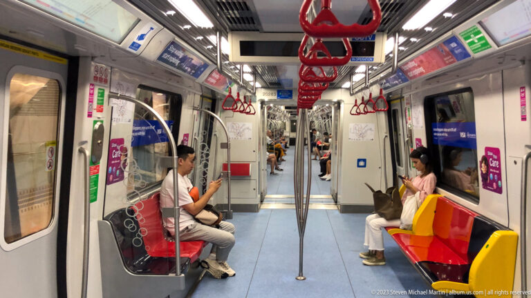 Passengers seated inside a Singapore MRT train with red straps and colorful seats.
