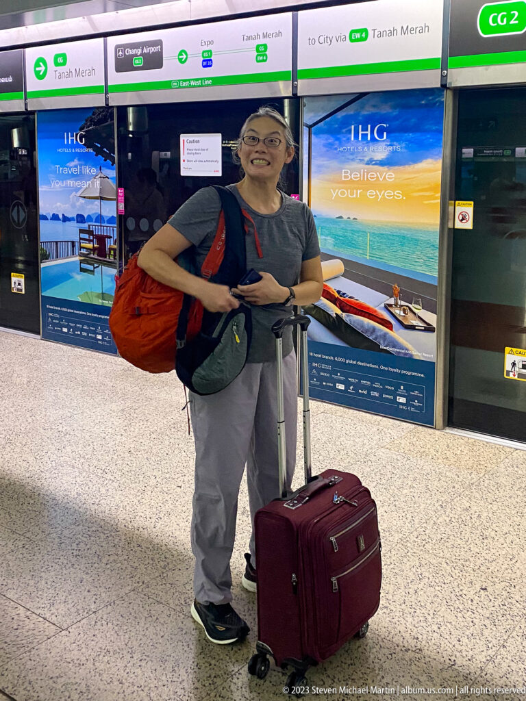 Female traveler standing with wheeled luggage at a Singapore MRT platform.