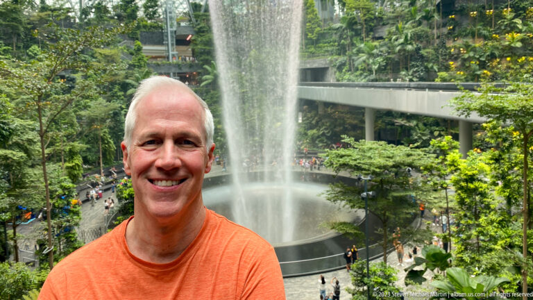 Smiling adult man in orange shirt standing before the Rain Vortex at Jewel Changi Airport.