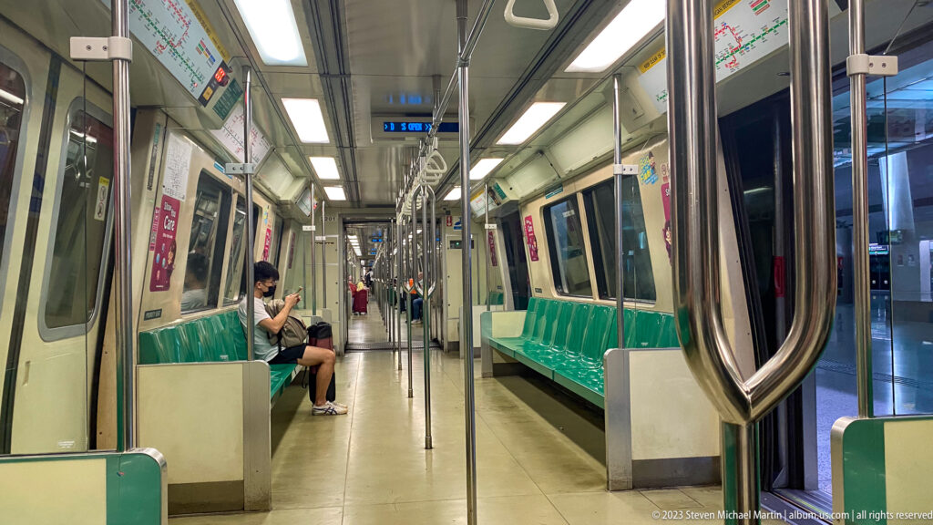 Interior of a Singapore MRT train with green seats, poles, handholds, and one passenger.