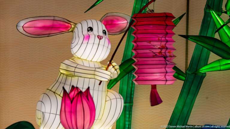White rabbit lantern with long ears holding a pink hanging lantern beside green bamboo forms.