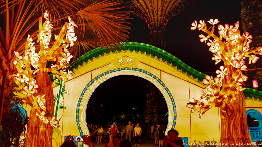 Circular green-roofed gateway between glowing blossom trees, with visitors and a Supertree in the background.