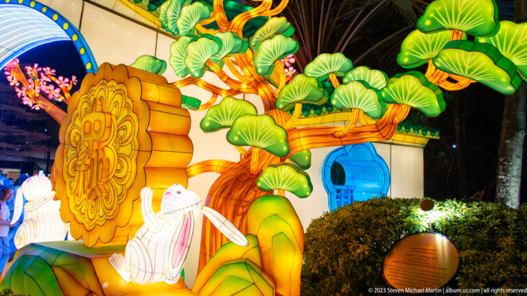 Side view of a golden mooncake lantern, white rabbits, green leaves, and an illuminated tree beside a gateway.