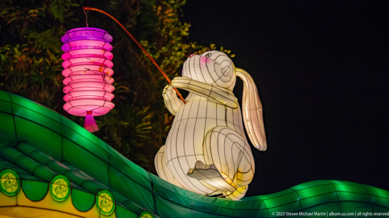White rabbit lantern seated on a curved green roof beside a hanging pink lantern at night.