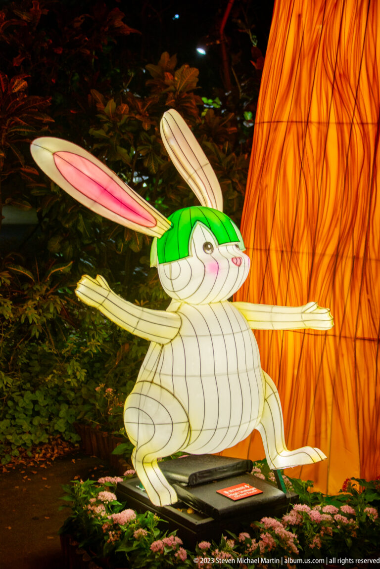 White rabbit lantern posed running with arms extended beside a glowing amber tree trunk.