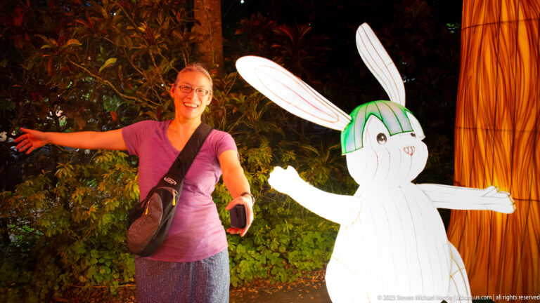 Adult woman posing with arms out beside a white rabbit lantern in a nighttime garden display.