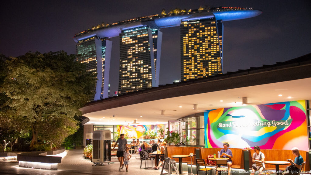 Open-air dining area with seated visitors and Marina Bay Sands illuminated behind it.