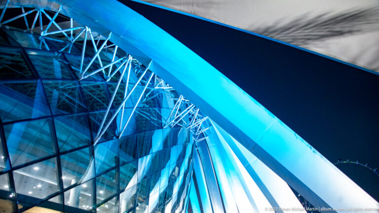 Blue-lit diagonal beam crossing a glass façade with exposed steel trusses.