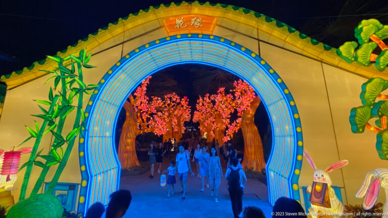 Full moon-gate façade with green roof, bamboo, rabbits, and blossom trees beyond.