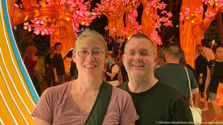 Two smiling adult visitors beneath glowing pink blossom trees in a crowded garden.