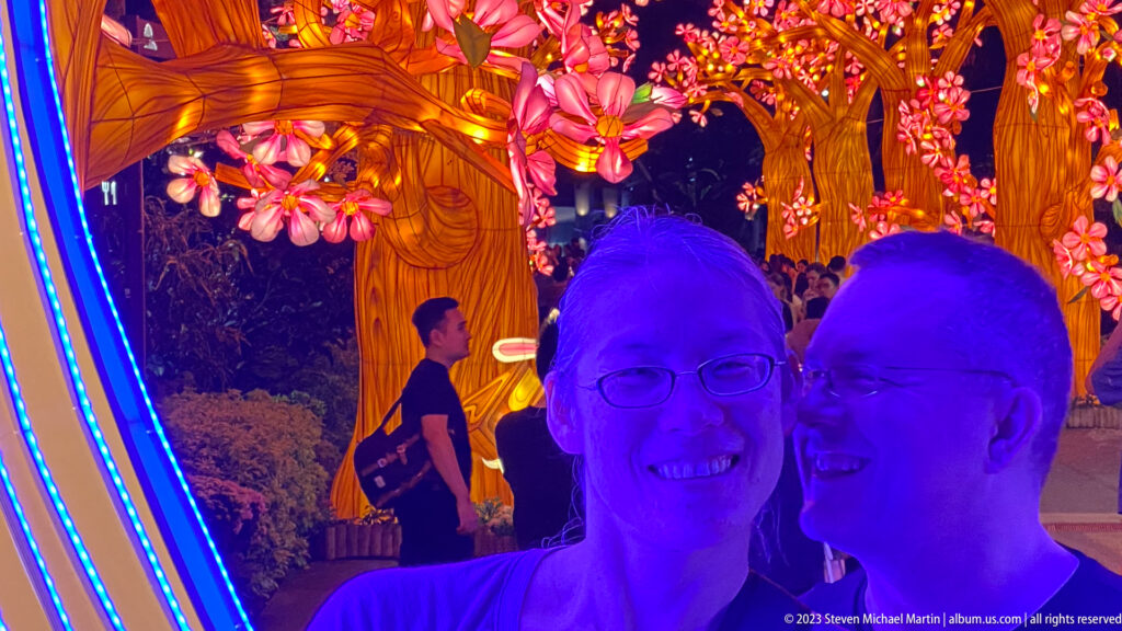 Two smiling adult visitors in blue light with amber blossom trees behind.