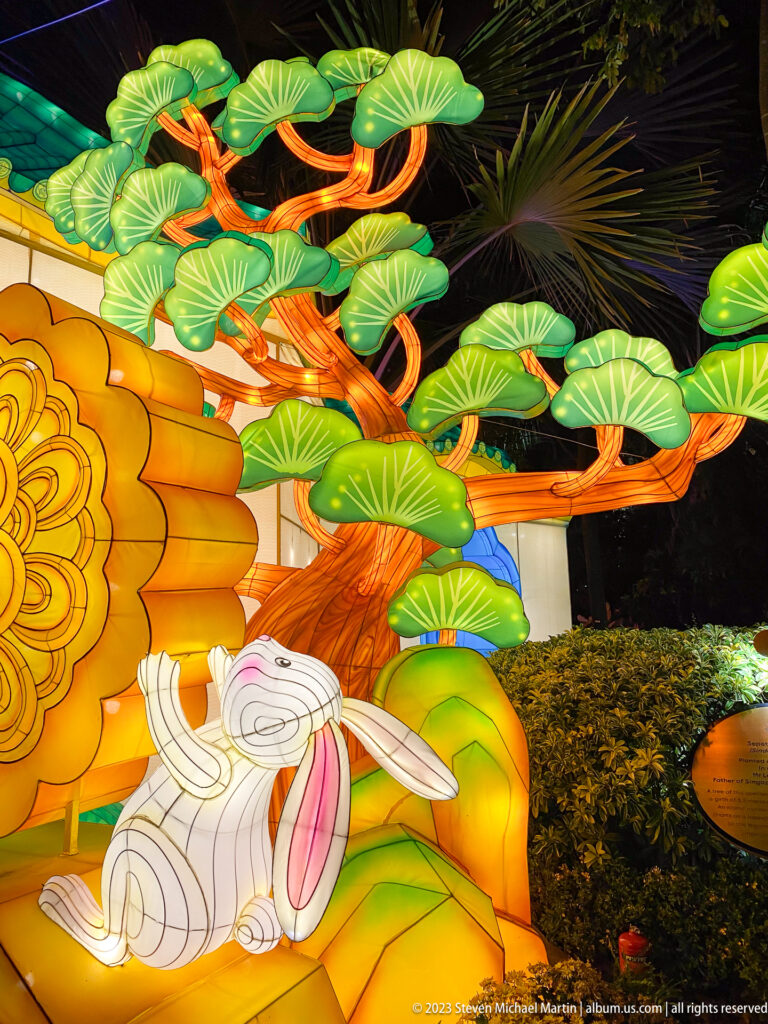White rabbit lantern beneath green leaves and orange branches beside a mooncake emblem.