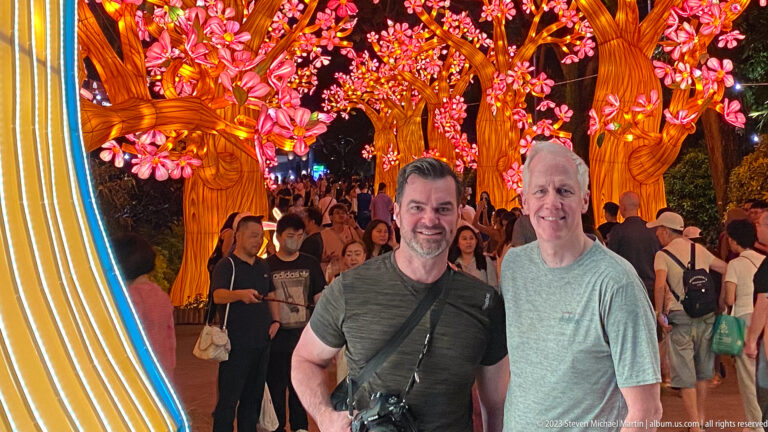 Two adult men posing with a camera beneath glowing blossom trees and a crowded walkway.