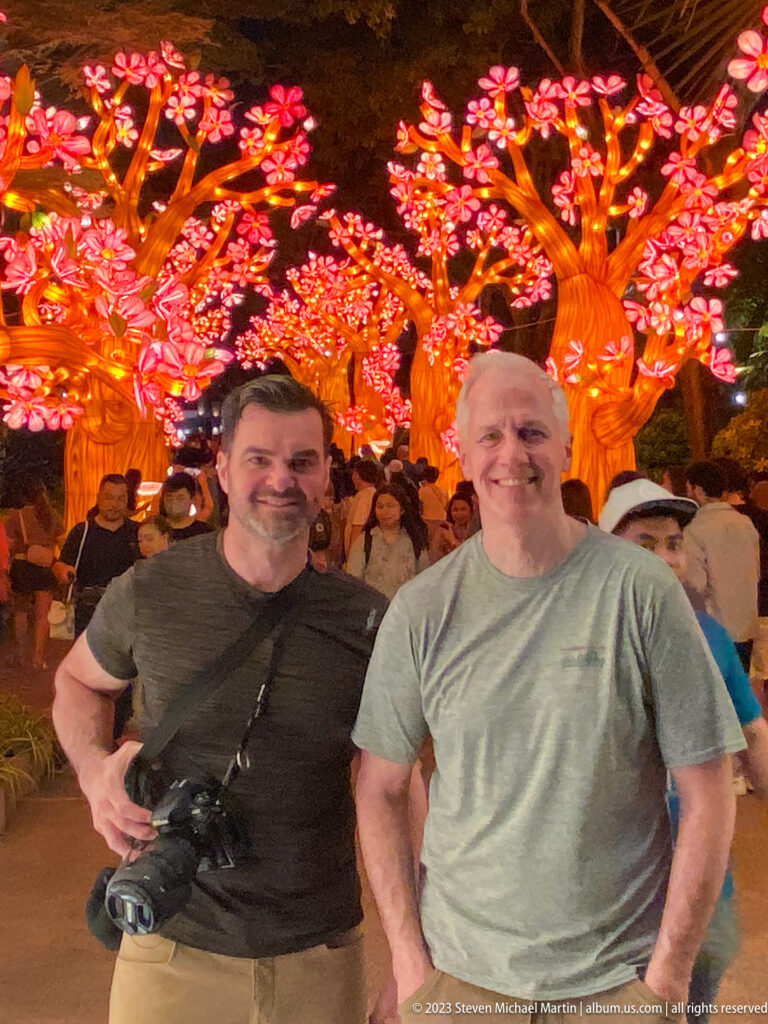 Two adult men standing beneath illuminated blossom trees amid a nighttime crowd.