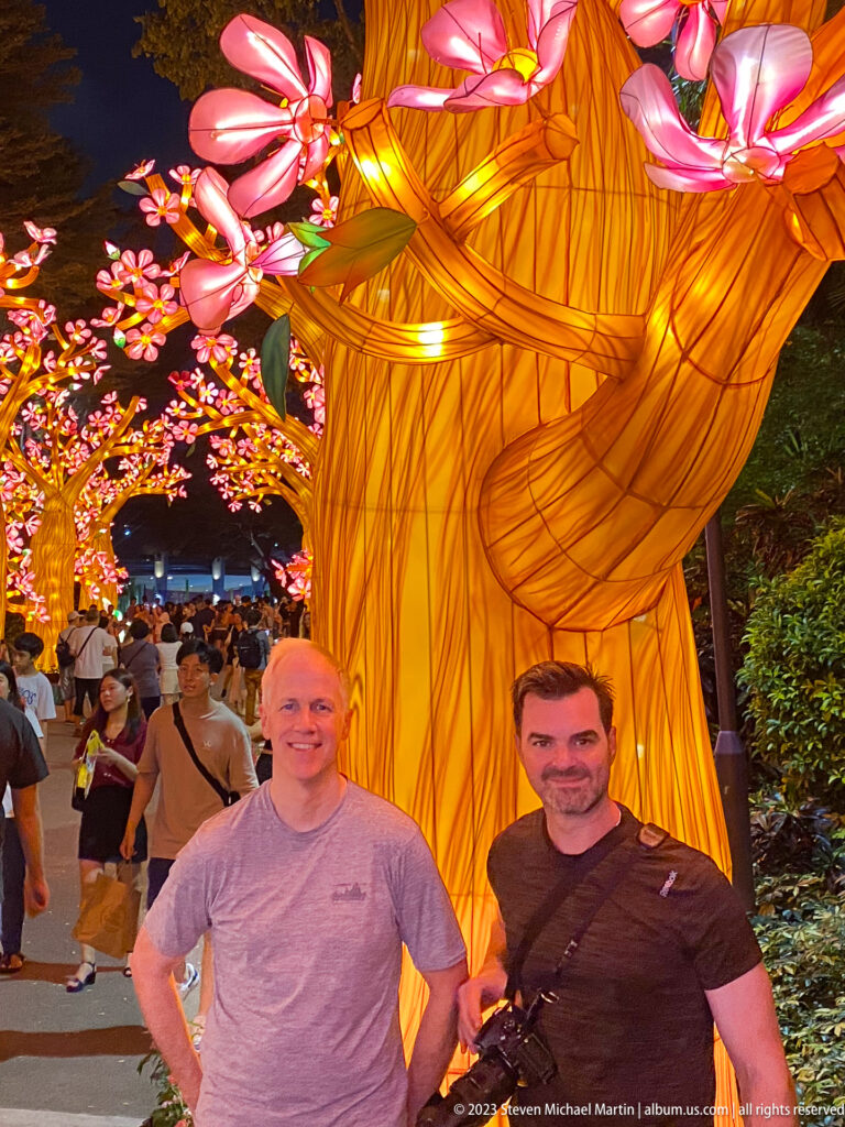 Two adult men posing beside a huge glowing tree trunk with pink blossoms overhead.