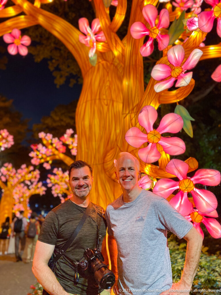 Two adult men posing beneath a glowing amber tree with large pink blossoms.