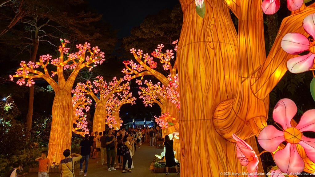 Large glowing tree trunk before a crowded walkway lined with blossom lanterns.