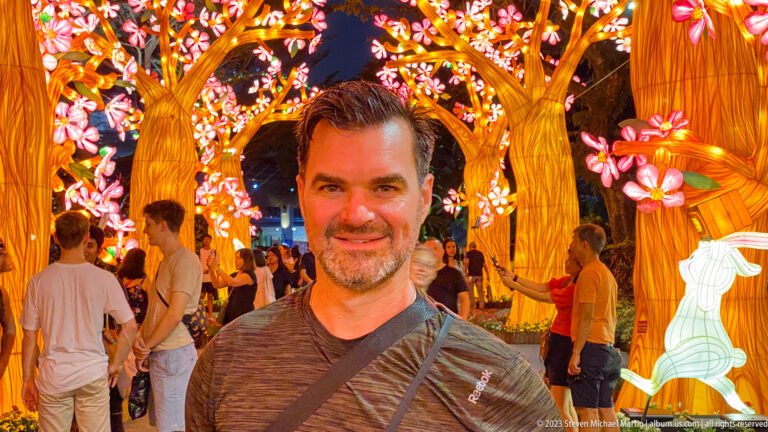 Smiling adult man beneath blossom trees with visitors and a rabbit lantern behind.