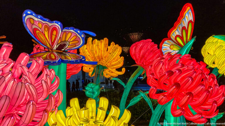 Colorful oversized flower lanterns with butterfly figures glowing against a dark nighttime garden.