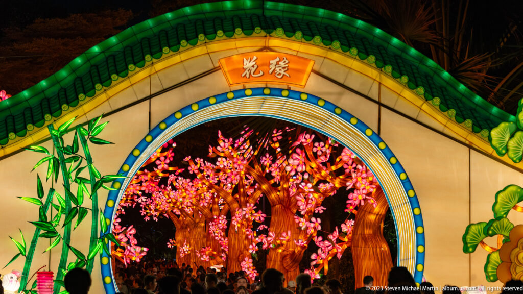 Illuminated Chinese-style moon gate with green roof, bamboo decoration, and pink blossom trees beyond.