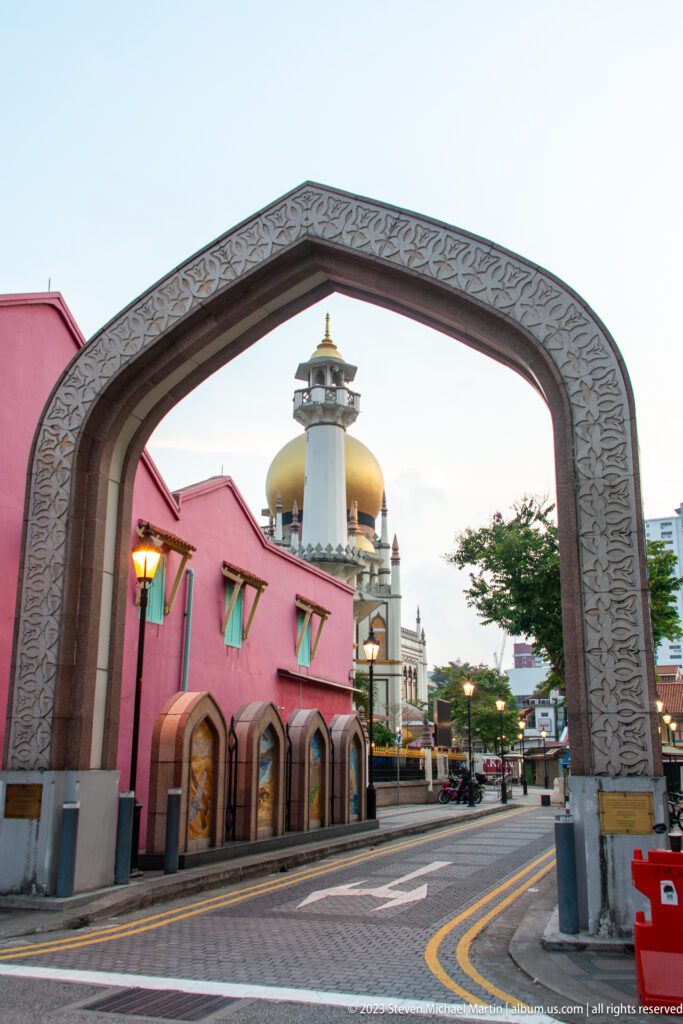 Ornamental pointed gateway framing Masjid Sultan’s golden dome and minaret along a curving city street.