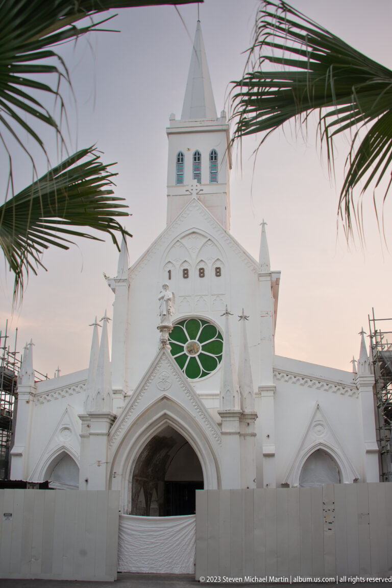 White Gothic Revival church façade with pointed arches, pinnacles, green tracery, palm fronds, and construction barriers.