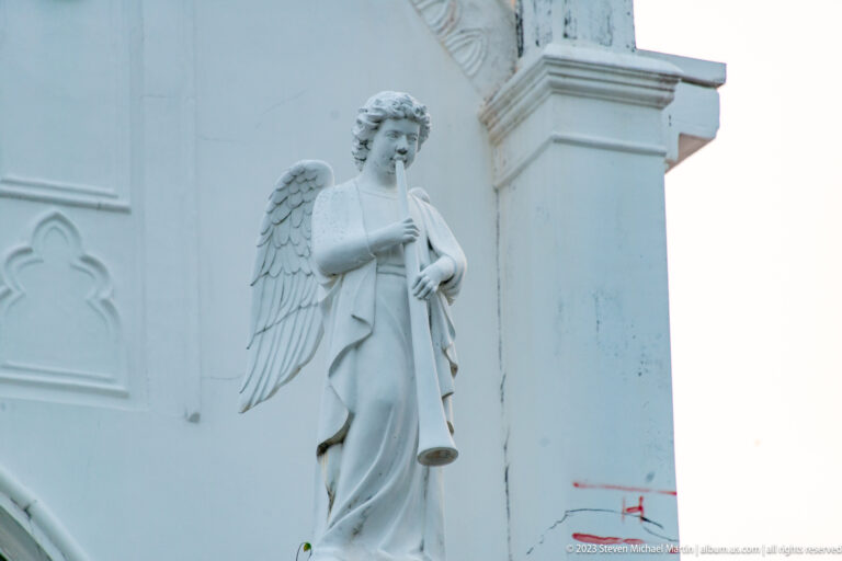Stone angel with wings holding a trumpet against a white church wall.