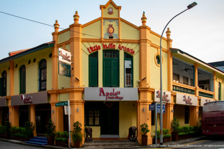 Little India Arcade corner building with clock pediment, green shutters, verandas, shop signs, and street traffic.