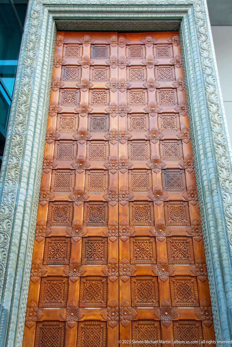 Large carved wooden door with many square patterned panels in an ornate metallic-looking frame.