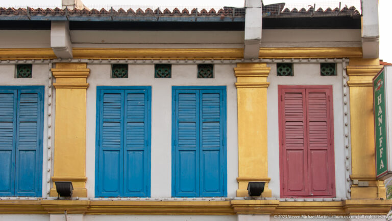 Yellow shophouse façade with blue and rose shutters, ochre pilasters, and green ventilation blocks.