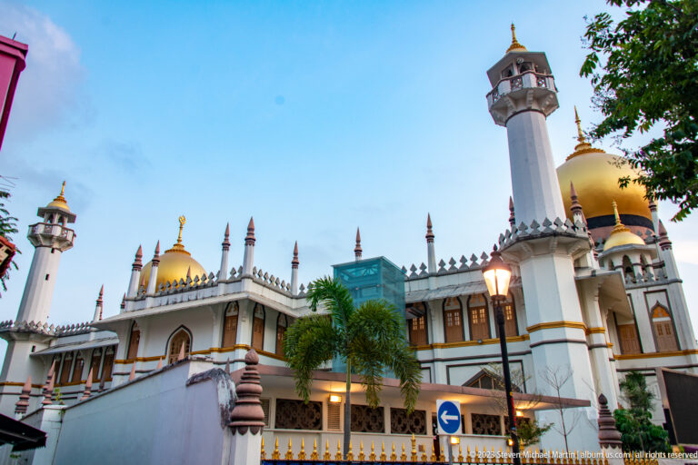 Low-angle view of Masjid Sultan with golden dome, minarets, finials, palms, and nearby buildings.