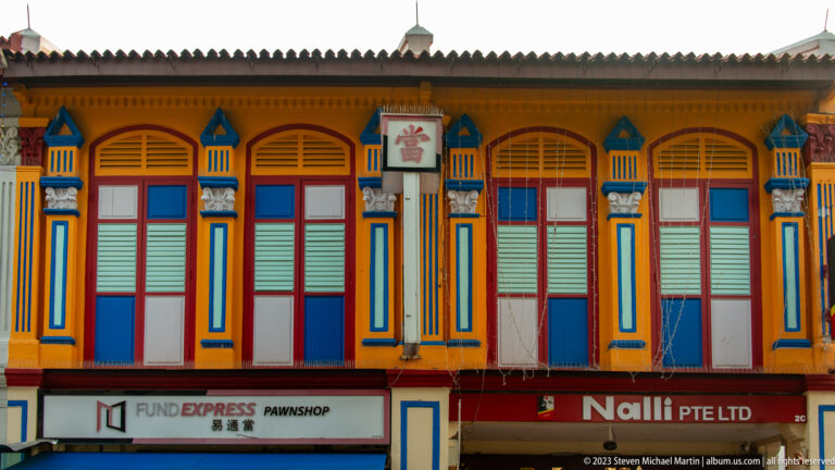 Colorful shophouse façade with ochre walls, blue pilasters, red frames, and green shutters.