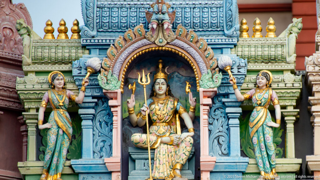 Murugan holding a spear between Valli and Deivanai in a colorful temple niche with peacock motifs.