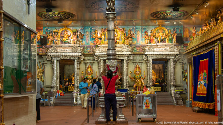 Temple interior with gilded shrines, many deity figures, hanging lamps, flagstaff, and worshippers.