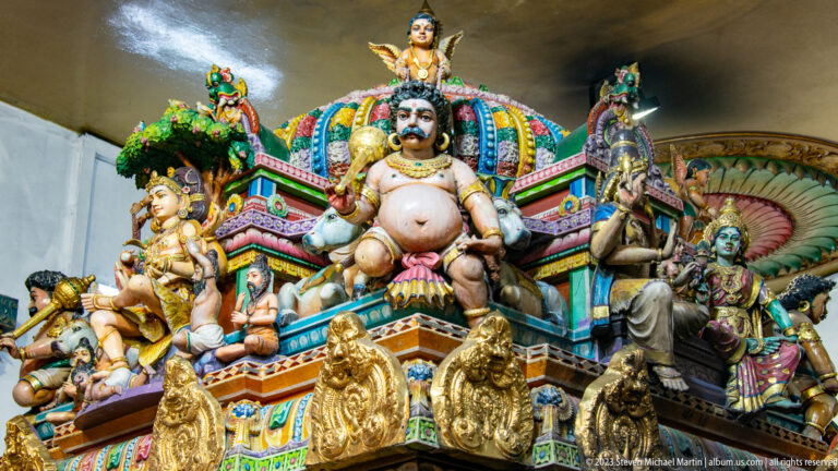 Colorful temple roof sculpture with central seated deity, attendants, animals, and ornate canopy.