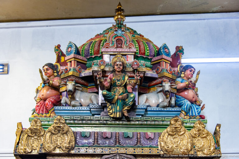 Seated goddess beneath a colorful canopy, flanked by two elephants and guardian figures.