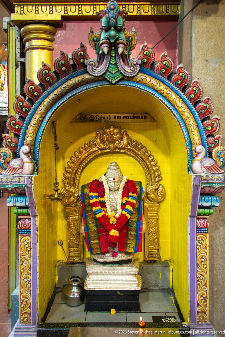 Garlanded Shiva lingam inside a yellow shrine niche with gilded arch and metal offering vessels.