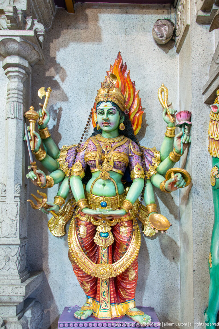Green-skinned multi-armed goddess with flaming crown, lotus, vessels, and gold jewelry.