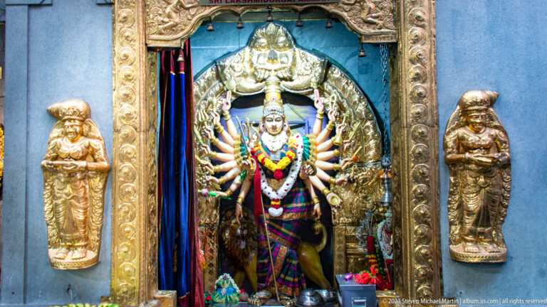 Garlanded multi-armed goddess in a gilded shrine with small attendant figures.