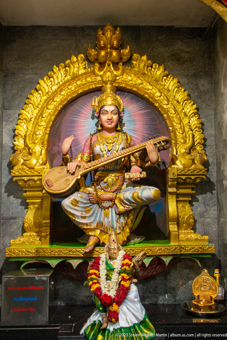 Saraswati seated with a veena beneath a gilded arch, with a white bird figure below.