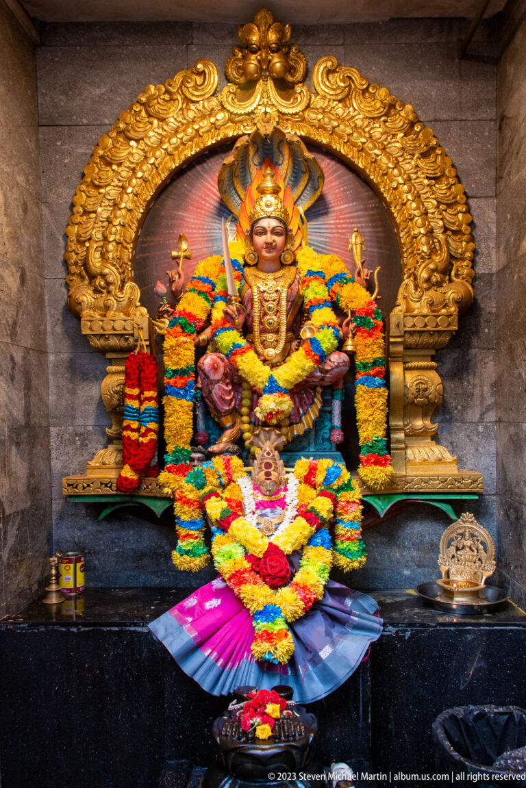 Seated crowned goddess beneath a gilded arch, surrounded by dense multicolored flower garlands.