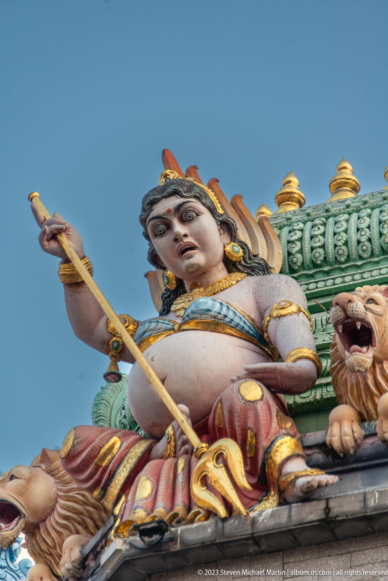Low-angle view of a painted male temple guardian holding a long staff against blue sky.
