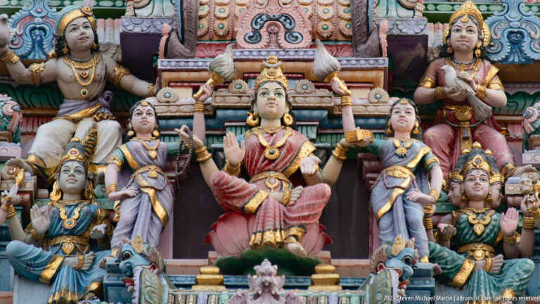 Seated crowned goddess in red surrounded by female attendants and smaller deities.
